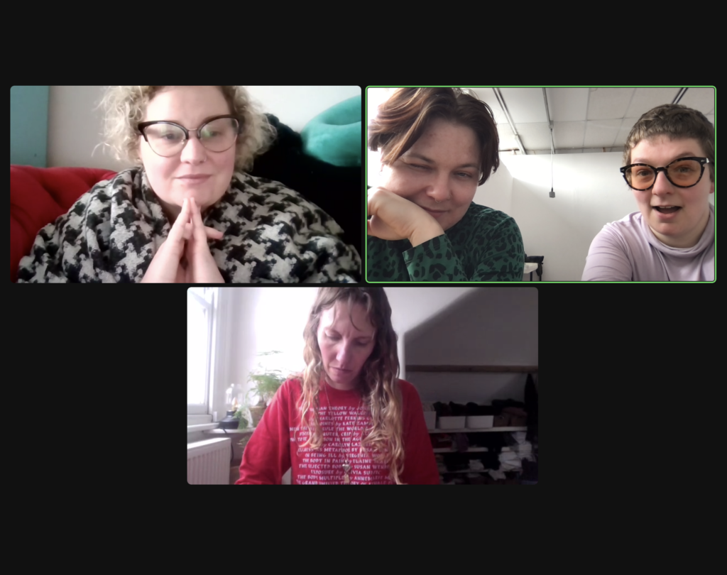 Screenshot from a Making Time zoom session showing three users on the call. One window shows Liza, a white person with curly blonde hair. Their fingers are steepled and they look pleased. Another window shows Jo, a white person with short brown hair, and Grace, a white woman with short brown hair and orange tinted glasses. Jo is winking and Grace is smiling. The last window shows Abby, a white person with long curly hair. Abby is looking down and is wearing a red shirt with the names of lots of disabled artists on it.